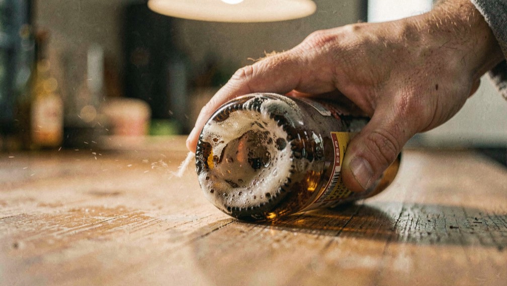 A hand rolling a brown beer bottle on its side across a wooden counter, cloudy sediment swirling in the last inch of beer, warm overhead light