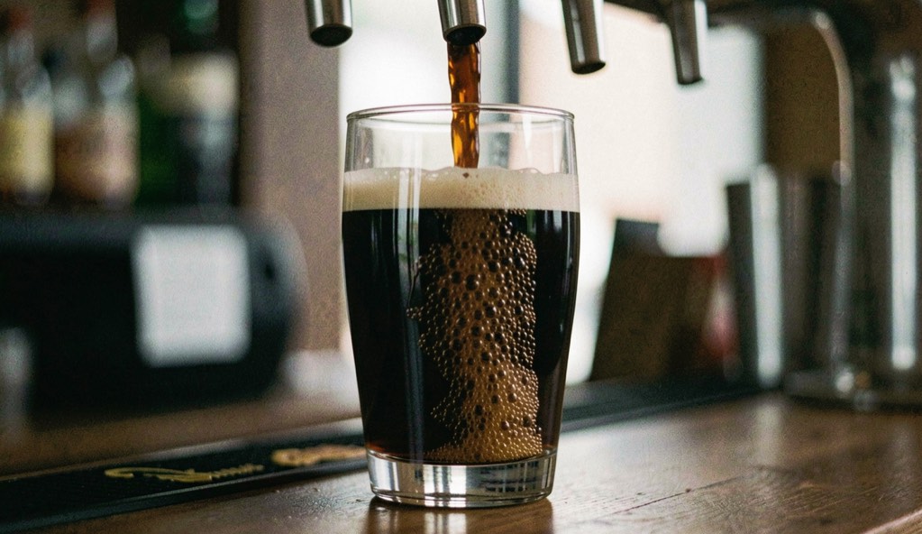 A dark beer cascading in a glass beneath a tap, the surge of fine bubbles moving downward through the liquid, bar light behind the glass