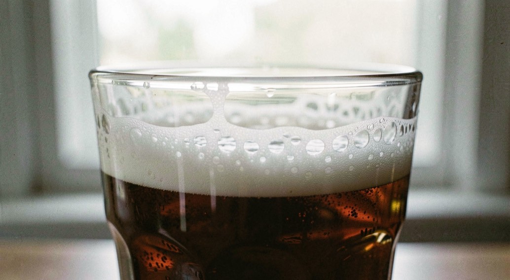 Close up of a pint glass with a dense white head, lacing rings down the inside of the glass, bright window light behind