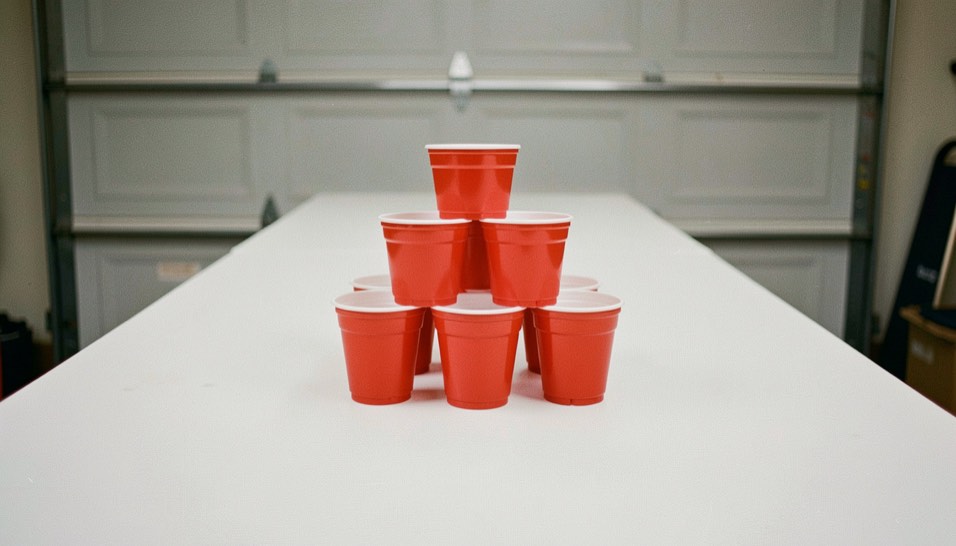 Ten red plastic cups racked in a tight triangle at the end of a long white table, apex pointing toward the camera, garage wall out of focus behind